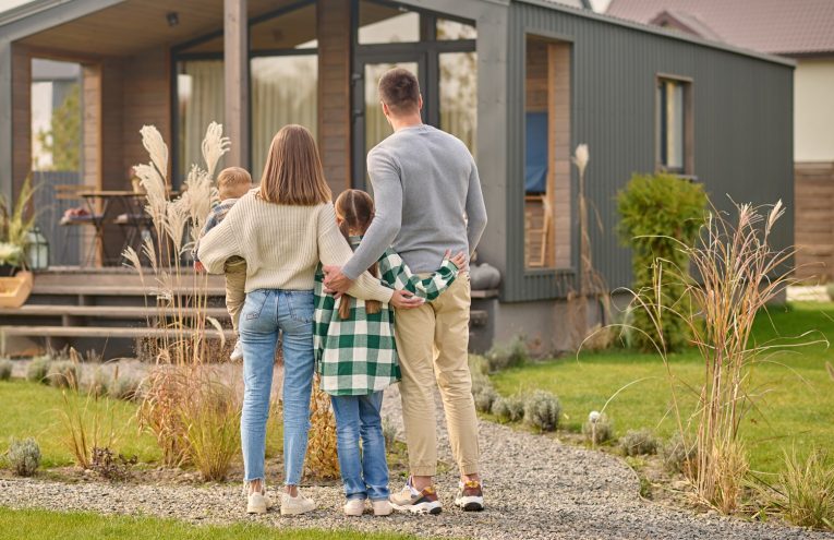 Happy family. View from back of man woman with child and girl standing together hugging and admiring their cozy home on fine autumn day